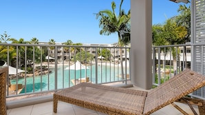A balcony with a wicker lounge chair, a pool, and a view of a resort area with palm trees and a clear sky. - The Sand Resort Kingscliff - Dual key 2B (6217/18) (Casuarina)