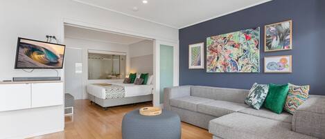 A modern living room with a grey sofa, a grey ottoman, a flat-screen TV, and a bed with a mirror.