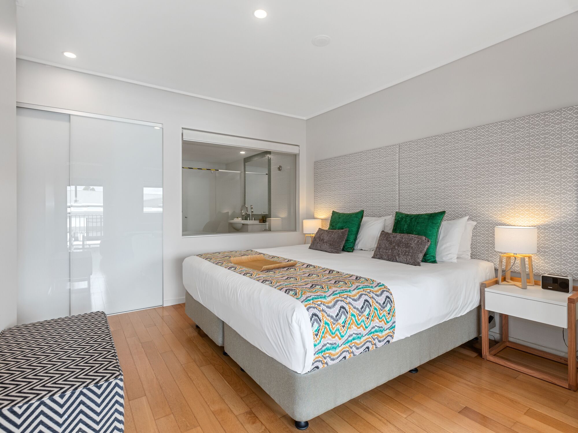 A modern bedroom with a large bed, a bedside table with a lamp, a mirror, and a patterned wall.