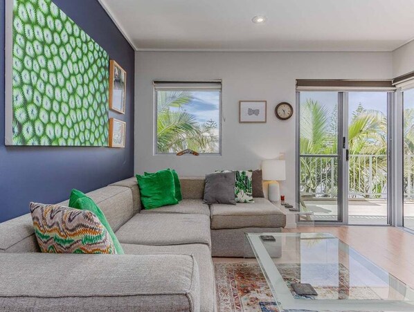 A modern living room with a sofa, a glass coffee table, and a view of palm trees through large windows.