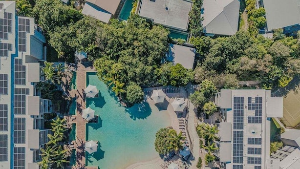 Ovenudblik over et hotel med en central swimmingpool omgivet af frodig vegetation og flere bygninger med solpaneler.