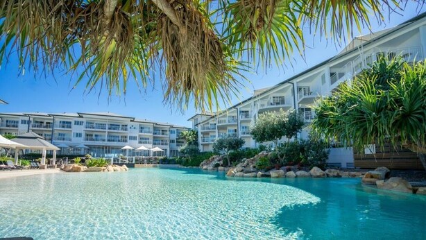 Una piscina cristalina con sillones de playa y sombrillas, rodeada por un edificio de resort con varios balcones.