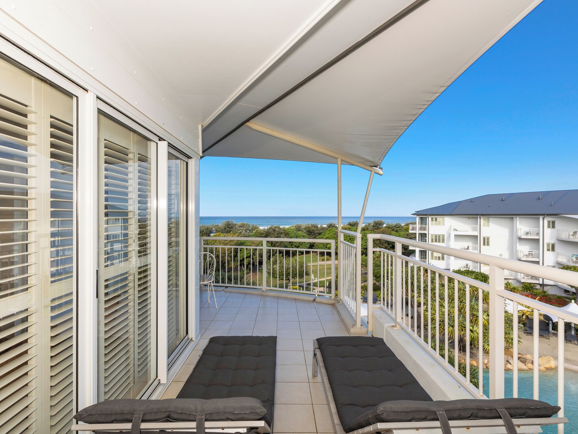 A balcony with two sunbeds, a white awning, and a view of the ocean and other buildings.