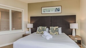 Una cama bien hecha con sábanas blancas, dos almohadas decorativas y una toalla enrollada al lado. Hay dos lámparas de noche sobre mesitas de madera, y un cuadro enmarcado cuelga sobre la cama.