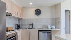 Una cocina moderna con armarios de madera clara, lavavajillas de acero inoxidable, cafetera negra y un reloj de pared.