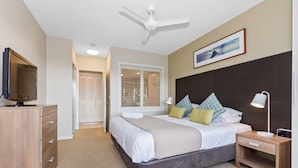 Une chambre à coucher comprenant un lit, une table de chevet, une commode, un ventilateur de plafond et un téléviseur posé sur une coiffeuse.