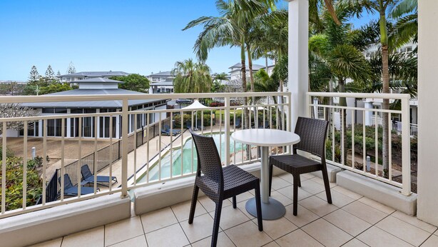 A balcony with two wicker chairs and a small round table, overlooking a swimming pool and palm trees. - The Pool Resort Kingscliff - 1 BR Small Pool View (7204) - Holiday Management Co. (Tweed)