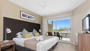 A hotel room with a large bed, a ceiling fan, a TV, and a balcony with a view of trees and buildings. - The Pool Resort Kingscliff - 1 BR Small Pool View (7204) - Holiday Management Co. (Tweed)