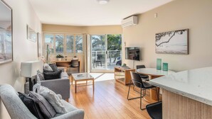 A spacious living area with a sofa, coffee table, and a dining table with chairs. There is a TV mounted on the wall and a balcony with a view of palm trees. - The Pool Resort Kingscliff - 1 BR Spa Suite (7314) - Holiday Management Co. (Tweed)