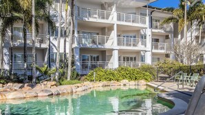 A pool area with lounge chairs, a clear pool, and a multi-story building with balconies. - The Pool Resort Kingscliff - 1 BR Spa Suite (7314) - Holiday Management Co. (Tweed)