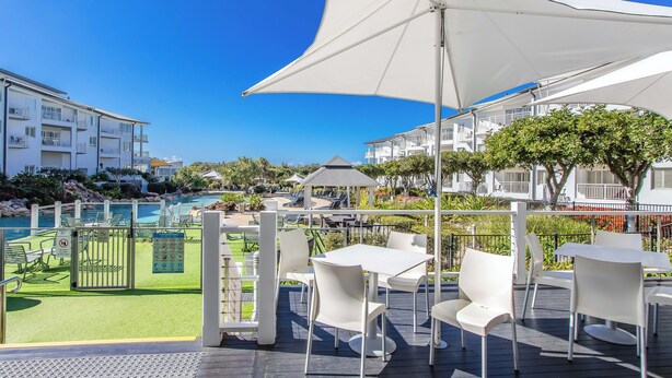 A poolside area with white tables and chairs, a large umbrella, and a view of a swimming pool and buildings in the background. - The Pool Resort Kingscliff - 1 BR Spa Suite (7314) - Holiday Management Co. (Tweed)