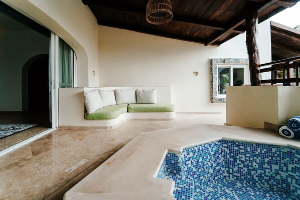 Zona de piscina con borde de baldosas, área de descanso acolchada con cojines y estructura de techo de madera.