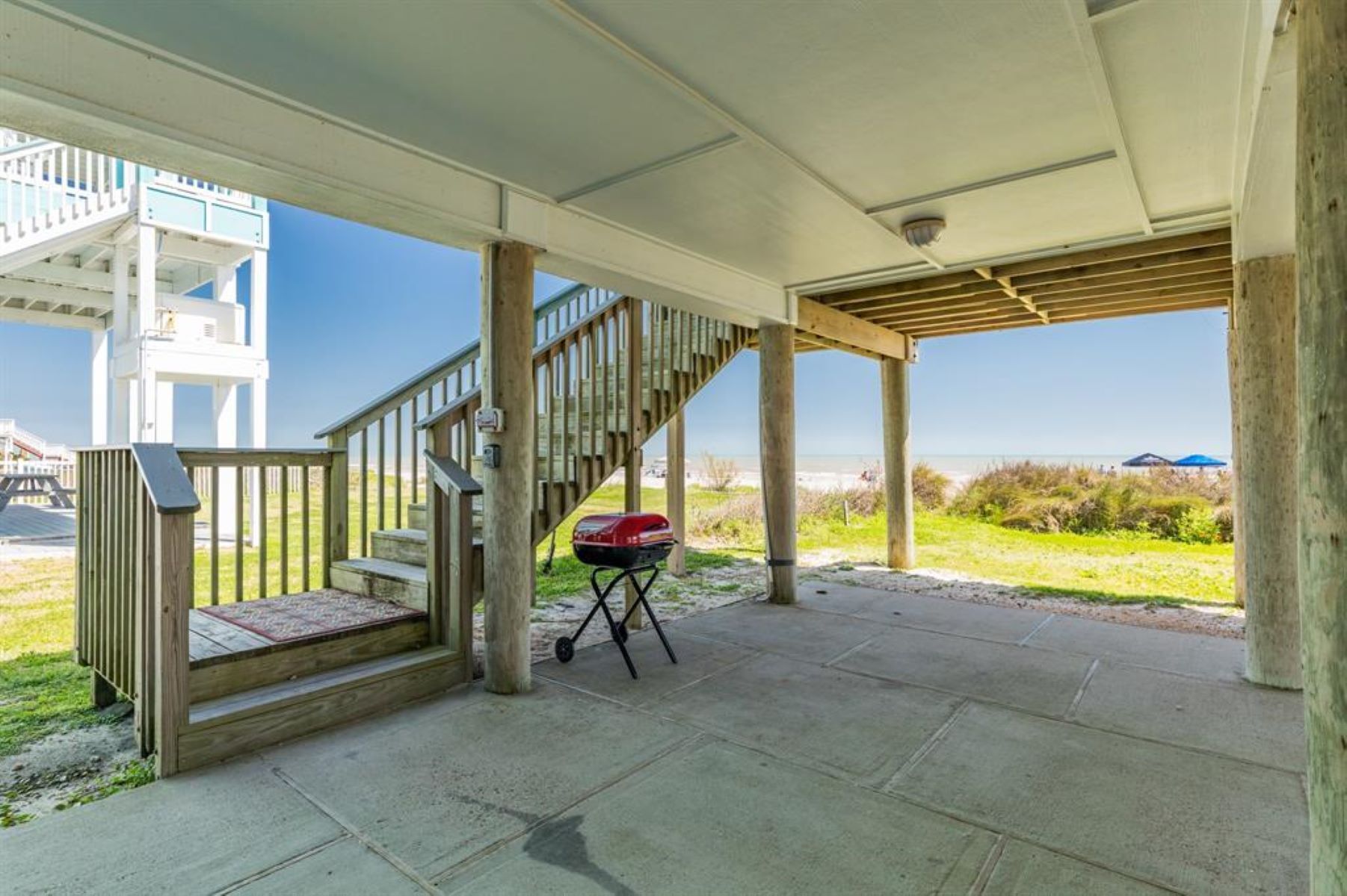 A covered outdoor area with a wooden staircase leading to a deck, a barbecue grill, and a view of the beach.