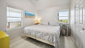 Un dormitorio con cama, dos ventanas, ventilador de techo y mesita de noche con lámpara.