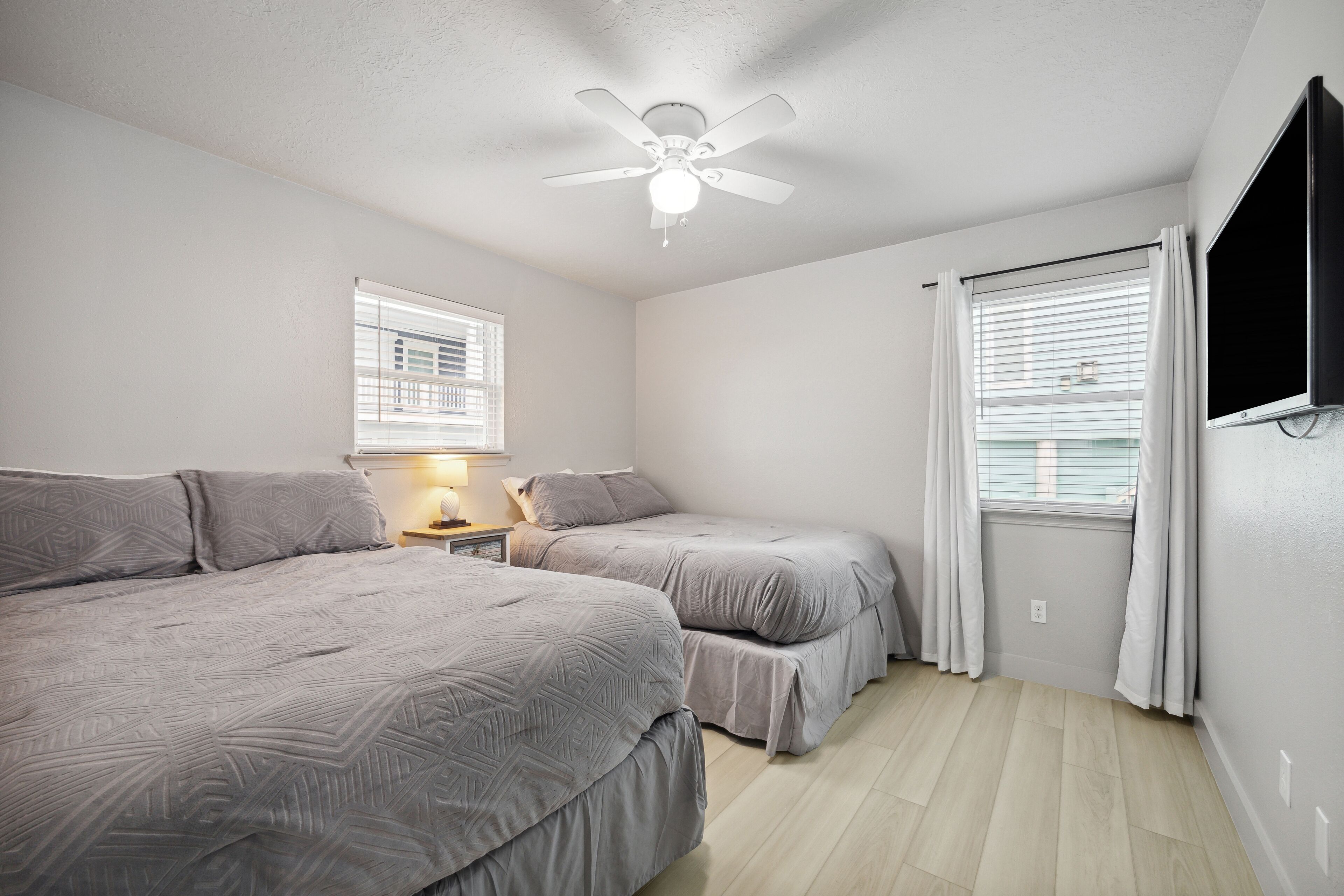 A bedroom with two beds, a ceiling fan, a window with blinds, and a TV mounted on the wall.