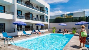 A pool area with lounge chairs and umbrellas, a multi-story building with balconies, and a clear blue sky. - Appartement au calme, à 15 min de la plage ! (La Rochelle)