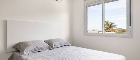 A bedroom with a bed, two pillows, a window with a view, and an air conditioning unit.