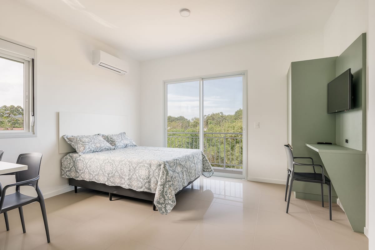 A bedroom with a bed, a desk, a chair, a TV, and a balcony with a view.