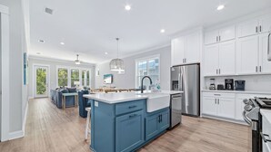 A modern kitchen with white cabinets, a blue island, stainless steel appliances, and a dining area with blue chairs. - Coastal Luxury Beach Escape with Plenty of Room! Pool and Golf Nearby (Orange Beach)