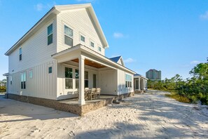 A large, single-story house with a covered porch and a small deck, situated on sandy ground with a clear sky. - Coastal Luxury Beach Escape with Plenty of Room! Pool and Golf Nearby (Orange Beach)