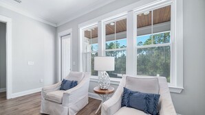 A room with two white armchairs, a small wooden table with a lamp, and large windows with a view of trees. - Coastal Luxury Beach Escape with Plenty of Room! Pool and Golf Nearby (Orange Beach)