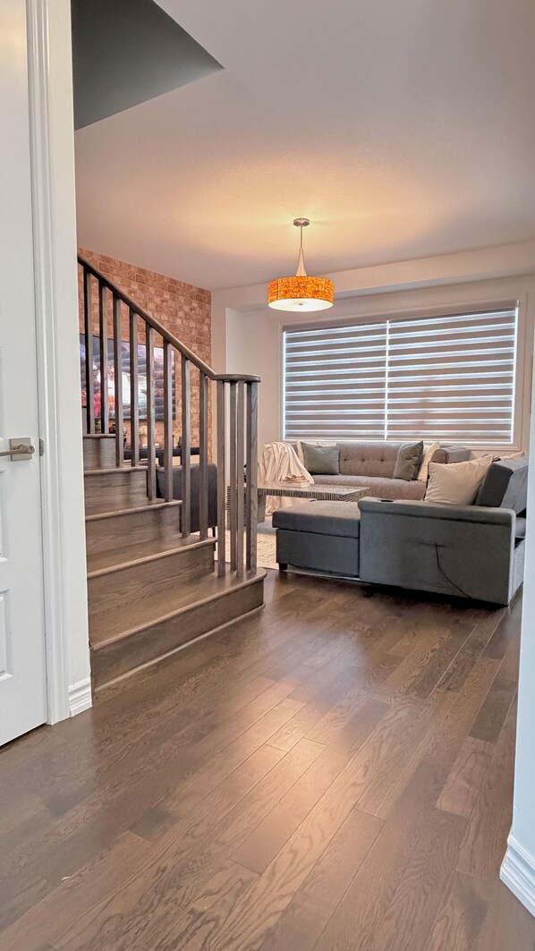 A modern living room with a staircase, a sofa, and a large window with blinds.