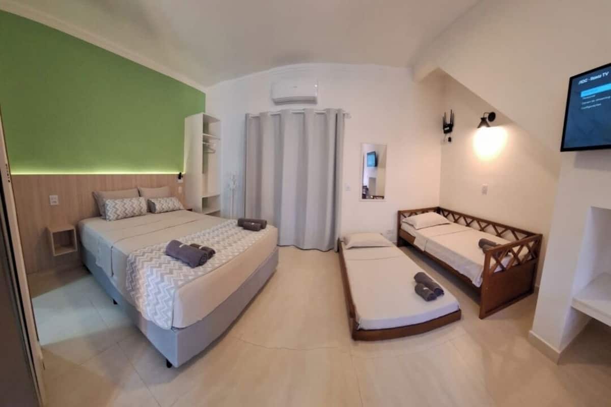 A compact hotel room with a large bed, a smaller bed, and a sofa bed. There is a TV mounted on the wall and an air conditioning unit above the sofa bed.