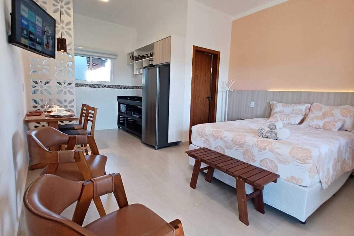 A modern hotel room with a large bed, wooden chairs, a small table, a kitchen area, and a TV mounted on the wall.