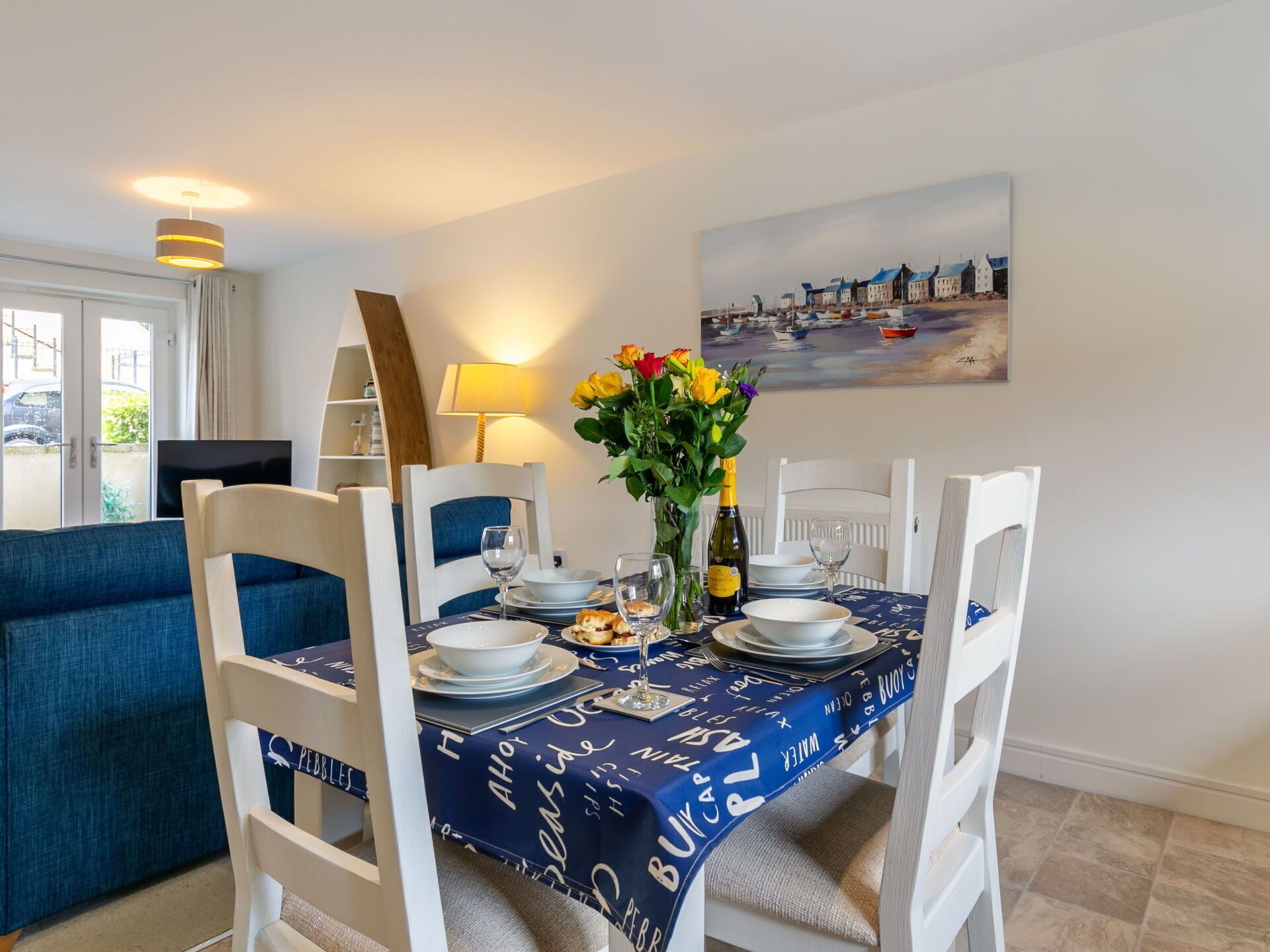 A dining area with a table set for four, white chairs, a vase of flowers, and a painting on the wall.