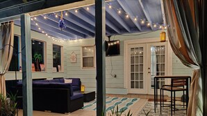 A covered patio area with a sofa, a table, and chairs. There are string lights hanging from the ceiling, and a television mounted on the wall. - Beach Villa with Private Pool - Walk to beach and Island View Casino (Gulfport)