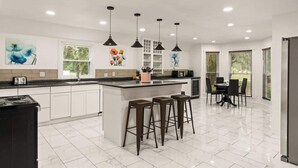 A modern kitchen with white cabinets, a central island with stools, pendant lighting, and artwork on the walls. - Poolside Oasis - Ask about snowbird specials! (Brunswick)