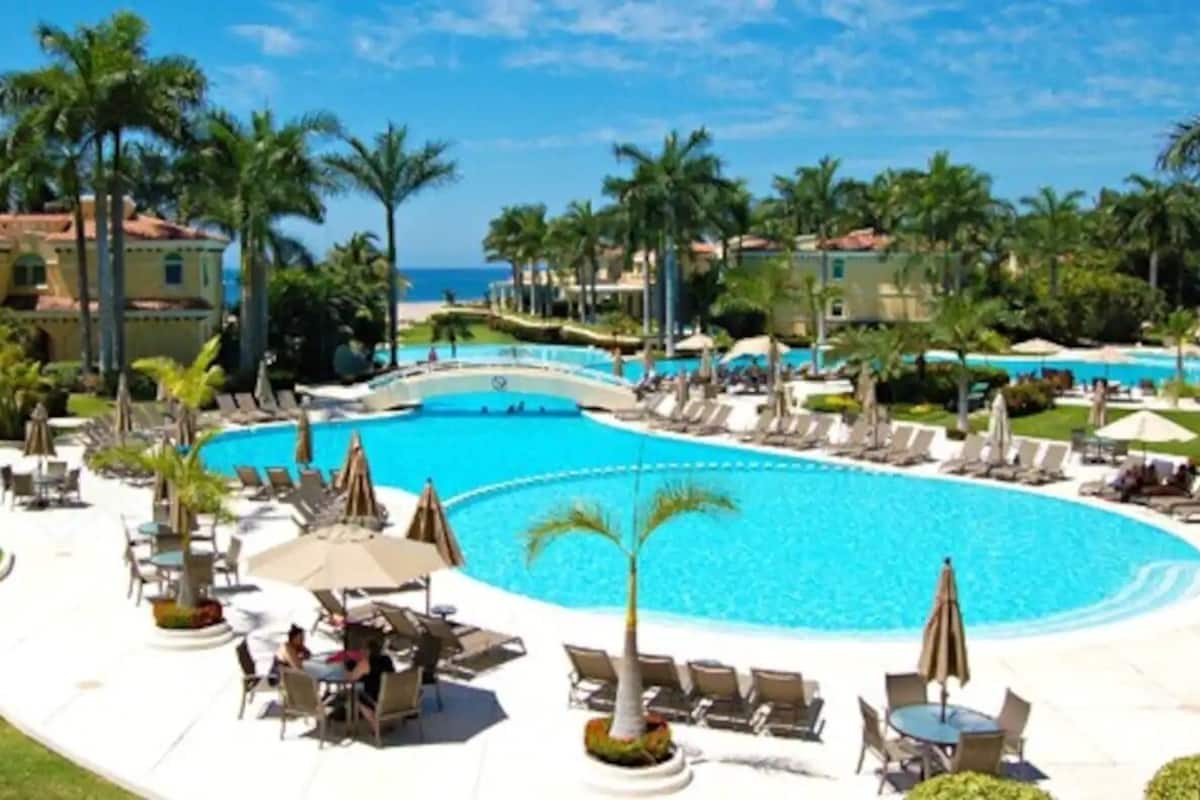 Un resort con una amplia piscina central, sillones de descanso, sombrillas y palmeras.