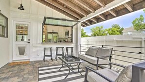 Un patio couvert aménagé avec des sièges, une cuisine visible à travers une porte vitrée et une vue sur l’extérieur.