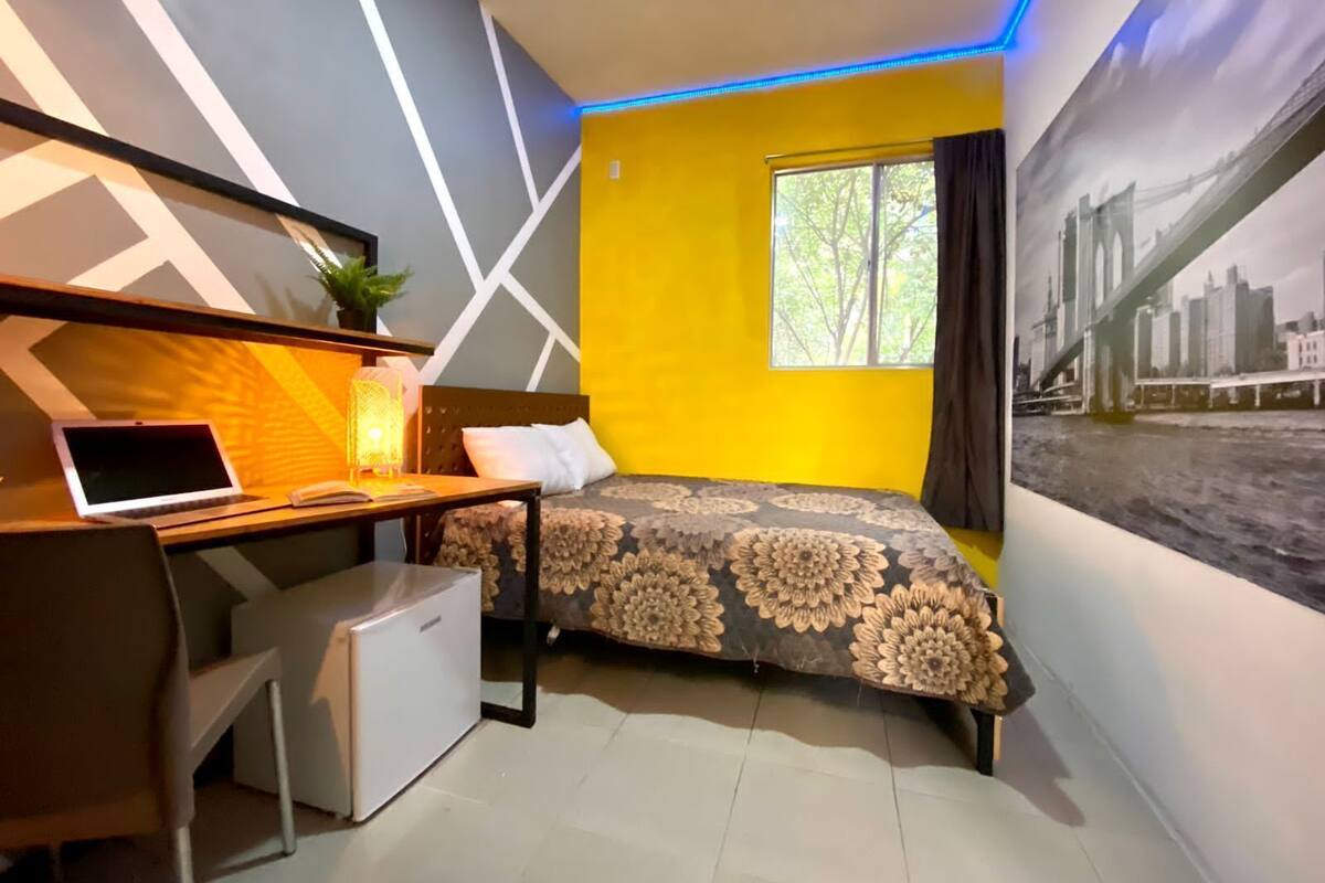 Una habitación moderna con cama, escritorio y silla, una ventana con cortinas y un cuadro de un paisaje urbano en la pared.