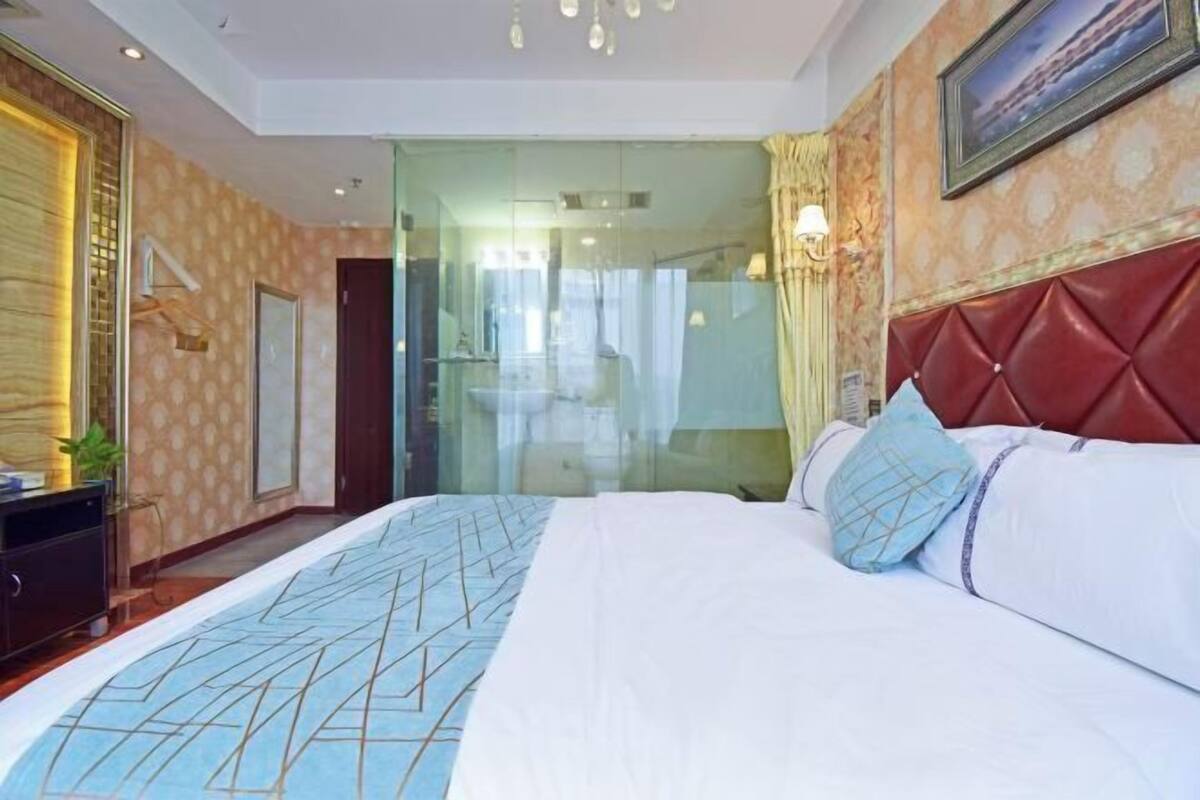 A hotel room with a bed, a glass partition, a red headboard, and a painting on the wall.