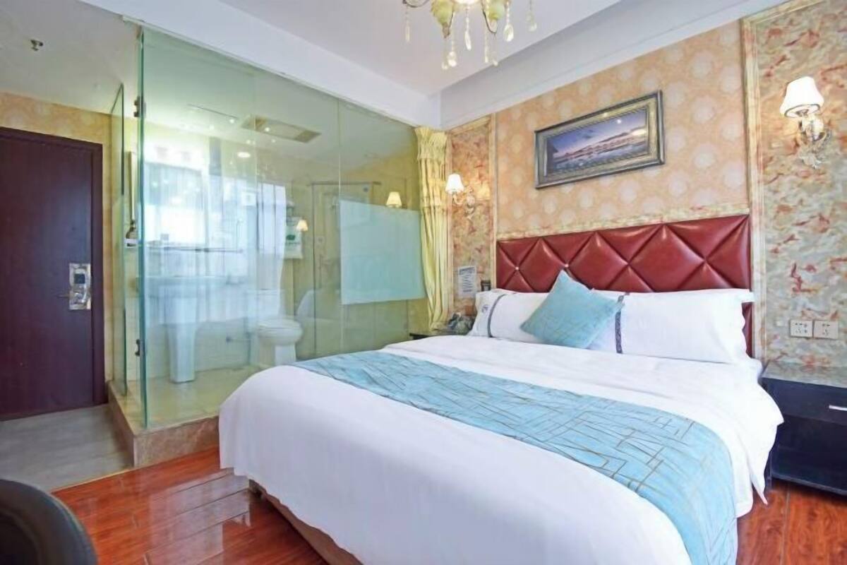 A hotel room with a bed, a glass shower enclosure, a wooden floor, and a wall-mounted light fixture.