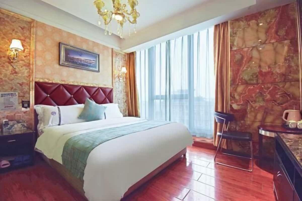 A hotel room with a large bed, a chandelier, a desk with a chair, and a view of the city through large windows.