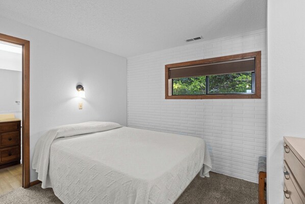 A bedroom with a bed, a window, a wall-mounted light, and a wooden dresser.