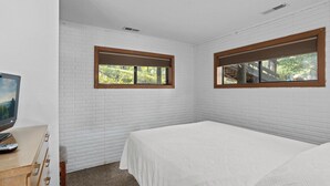 Makuuhuone, jonka seinät on valkoisella tiilellä vuorattu, ja jossa on valkoinen sängynpeite, puinen lipasto ja kaksi puukarmein varustettua ikkunaa.