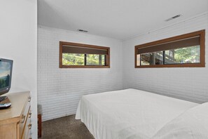 Una camera da letto con pareti in mattoni bianchi, un letto con copripiumino bianco, un comò in legno e due finestre con stipiti in legno.