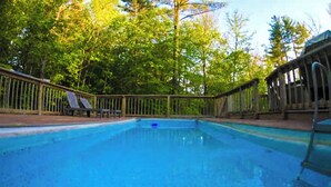 Una piscina con bordo in legno e sdraio, circondata da alberi.