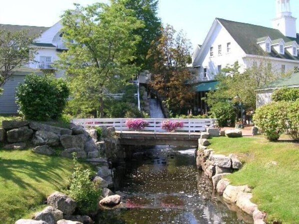 A bridge over a small stream with a white railing, surrounded by greenery and a building with a steeple in the background.