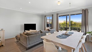 A modern living room with a grey sofa, a flat-screen TV, a dining table with white chairs, and large windows offering a view of greenery and a clear sky.