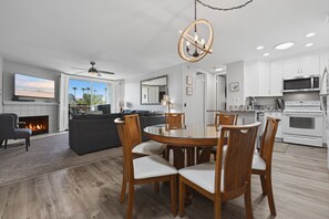 A modern living room with a fireplace, a dining area with wooden chairs, and a kitchen with white cabinets and appliances.