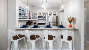 A modern kitchen with white cabinets, a stainless steel refrigerator, a microwave, and a coffee maker. There are four white bar stools with wooden seats. - Beach Blossom Villa - Privacy and Perfect Location (Fort Walton Beach)