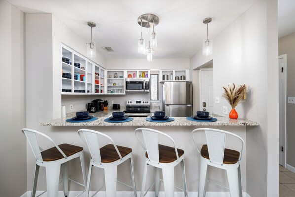 A modern kitchen with white cabinets, a stainless steel refrigerator, a microwave, and a coffee maker. There are four white bar stools with wooden seats.