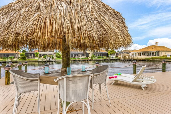 A wooden deck with a thatched umbrella, wicker chairs, and lounge chairs by a water body with buildings in the background.