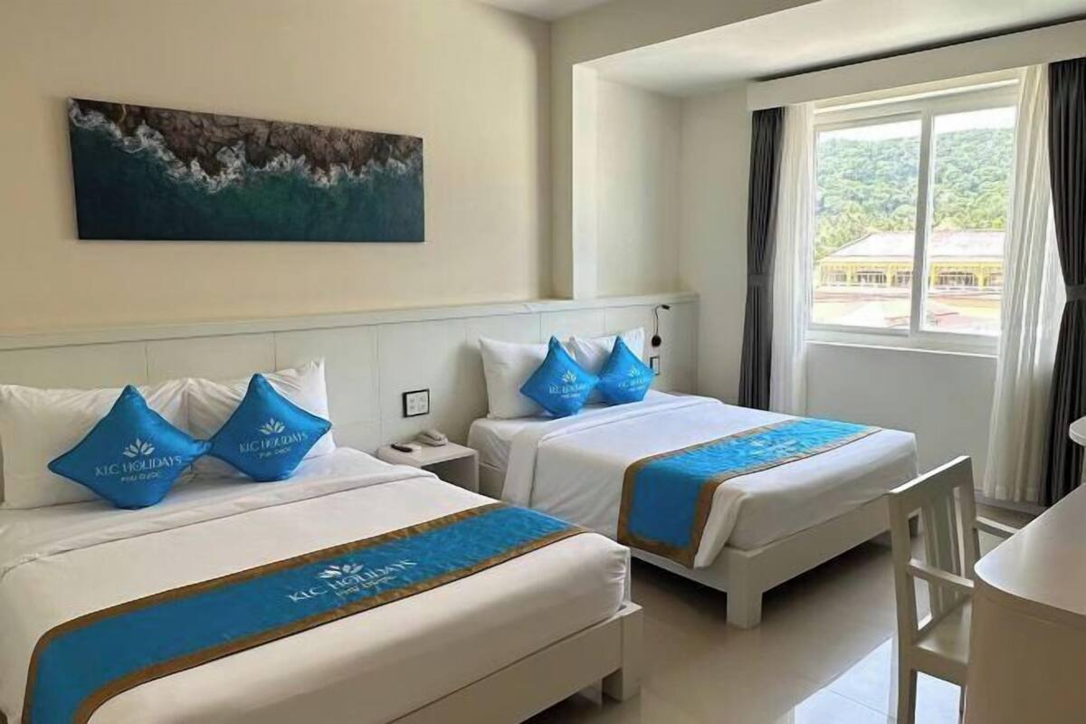 A hotel room with two beds, a desk, and a window with a view of greenery.