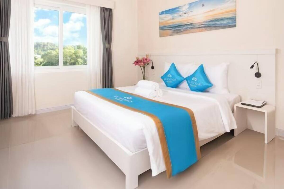 A hotel room with a bed, a nightstand, a window with curtains, and a painting on the wall.