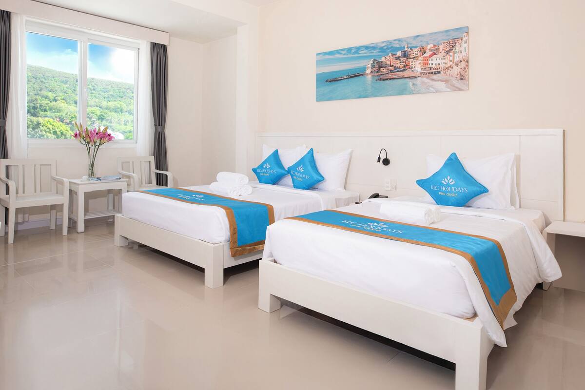 A hotel room with two beds, a desk, and a window with a view of a coastal town.